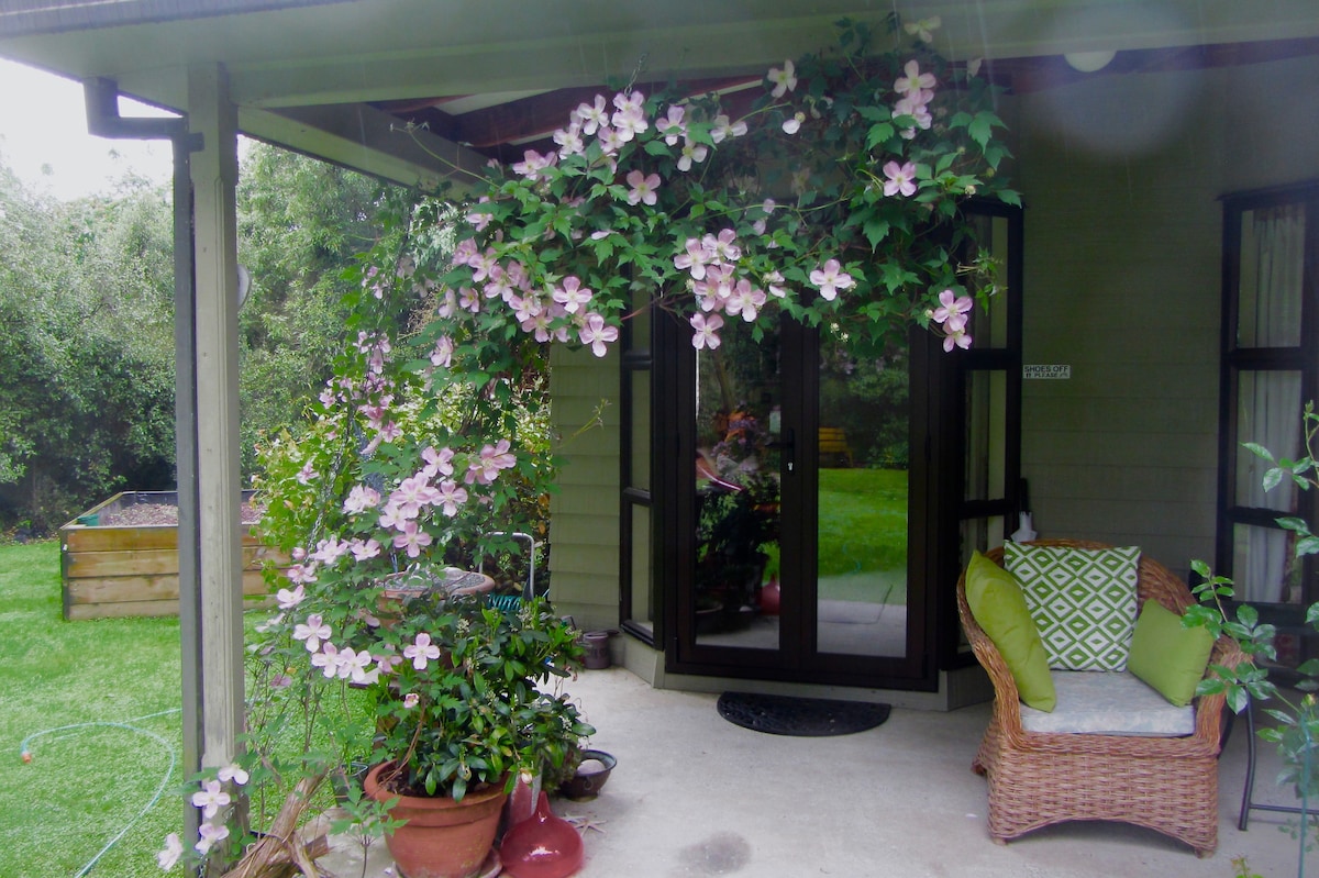 A welcoming entrance features a vibrant arrangement of pink flowers cascading from above. The doorway is framed by large glass doors, inviting natural light. A wicker chair adorned with green cushions is positioned beside potted plants, highlighting the lush garden surroundings.