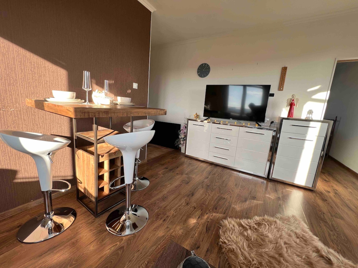 A modern living space is depicted with stylish bar stools and a high table for dining. A television is positioned on a white entertainment unit against the wall. Warm wood flooring complements the interior design, which is accentuated by natural light filtering in through the window.