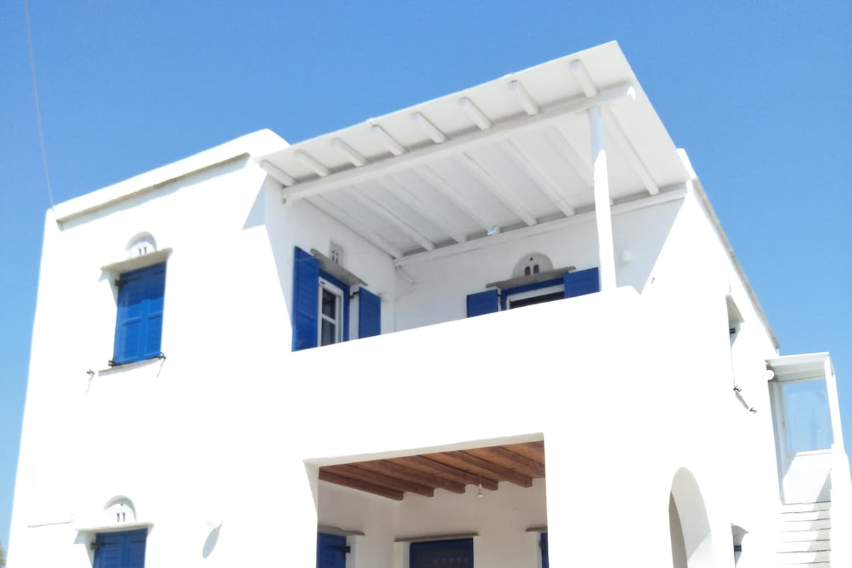 A white, two-story building features a spacious balcony with a pergola, offering a view of the blue sky. Blue shutters complement the white exterior, enhancing the overall coastal aesthetic. Architectural details, such as evenly spaced windows and a wooden ceiling, add to the structure's character.