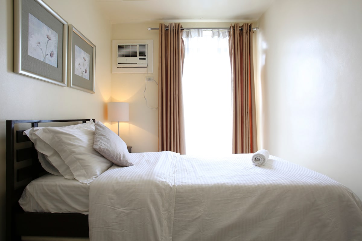 A cozy sleeping area features a comfortable bed with crisp white linens and decorative pillows. Natural light enters through a window dressed with sheer curtains, while framed artwork adorns the wall. An air conditioning unit is installed above the bed for climate control.