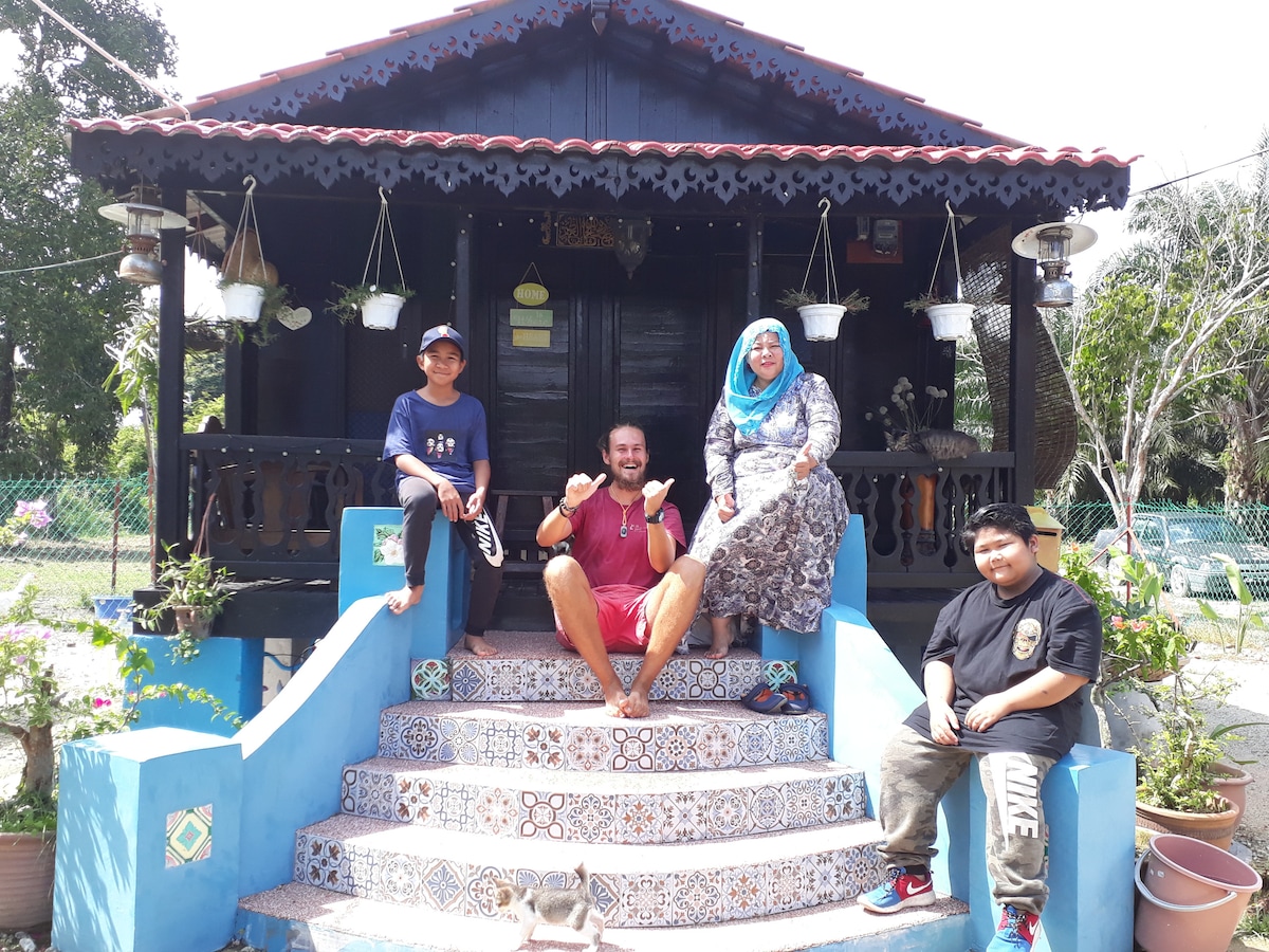 A traditional chalet with a blue and white tiled staircase is featured. Four individuals are seated on the steps, surrounded by decorative potted plants. The entrance is adorned with hanging pots, and a decorative railing adds to the rustic charm of the structure.