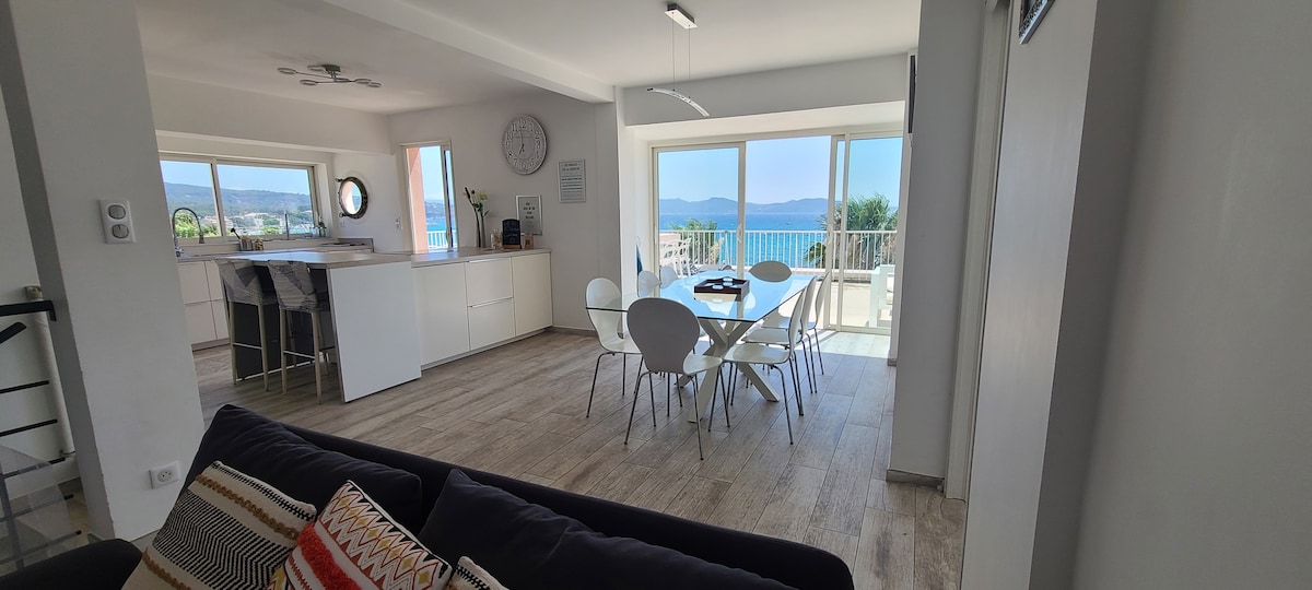 The spacious living area features modern furnishings and large glass doors, offering stunning views of the sea. A cozy sofa is positioned alongside a dining table, with a well-equipped kitchen visible in the background, illuminated by natural light.