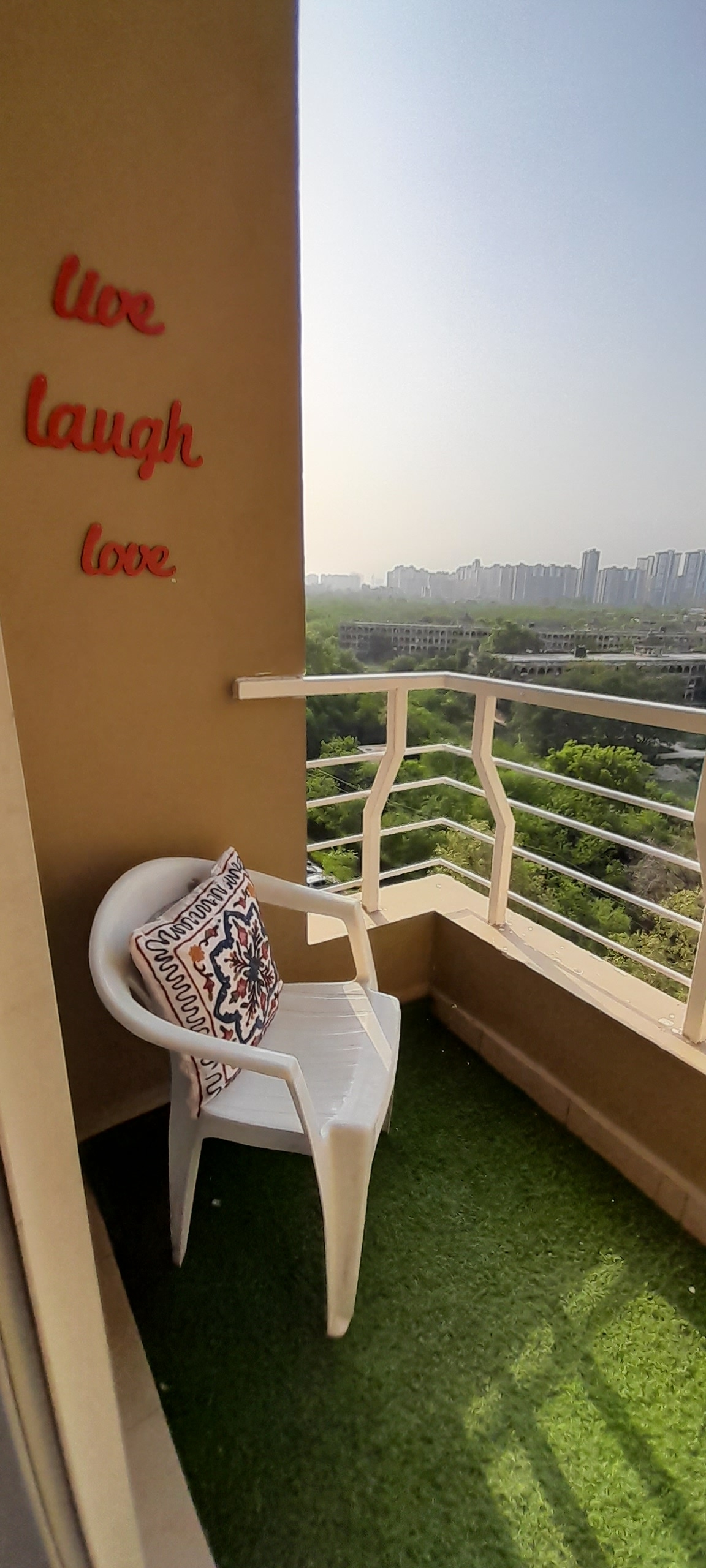 A balcony space is depicted with a white plastic chair and an accent pillow, positioned on green artificial grass. The adjoining wall features the words 'live laugh love' in a bold red font. A view of the surrounding greenery and city skyline is visible in the background.