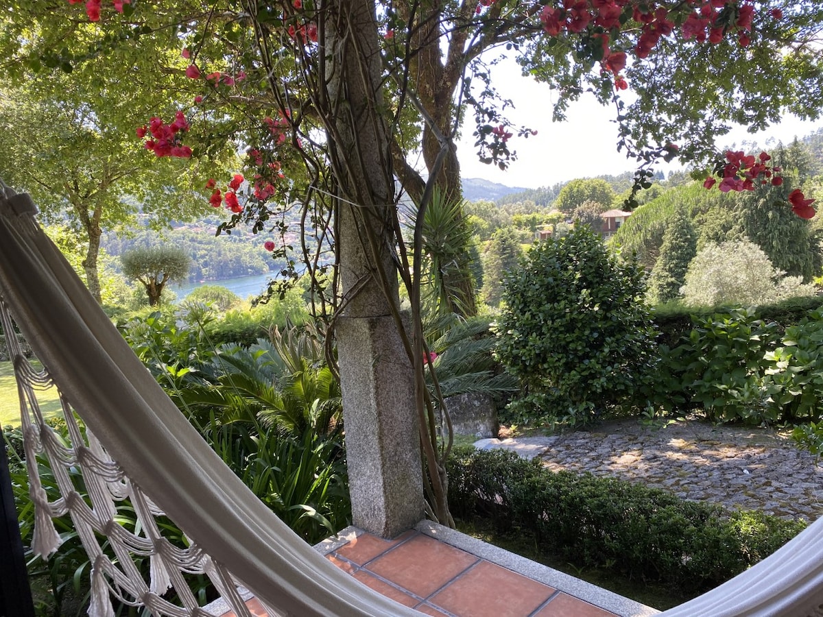 A serene view from a shaded porch features a comfortable hammock swaying gently. Lush greenery surrounds the space, providing a natural backdrop, while vibrant flowers add a touch of color. In the distance, a glimpse of the river can be seen through the trees.