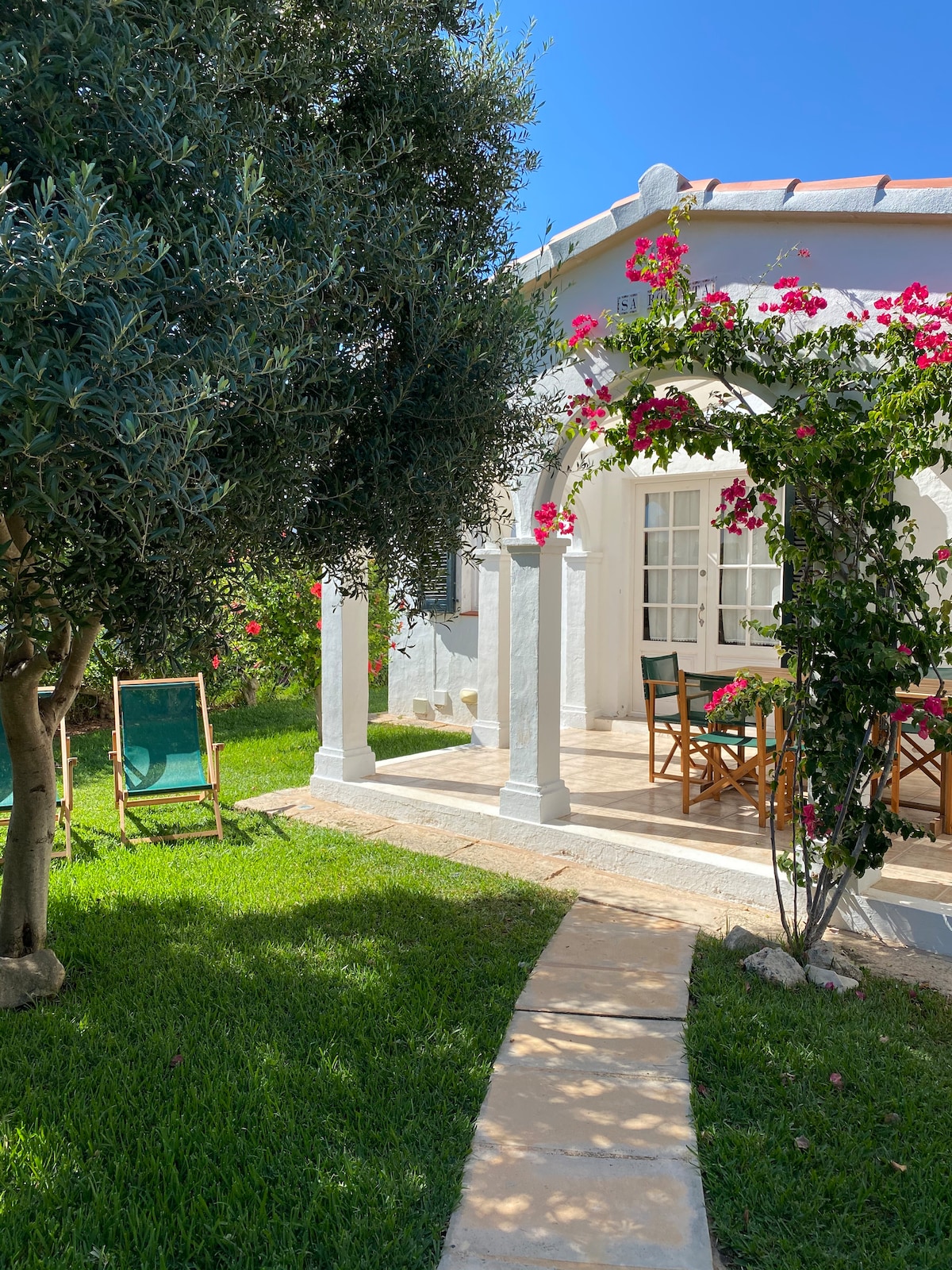 The image captures a charming outdoor space with vibrant greenery and flowering plants. A stone pathway leads to a covered porch featuring a dining table and chairs. A sun chair is positioned nearby, inviting relaxation in the serene garden setting.