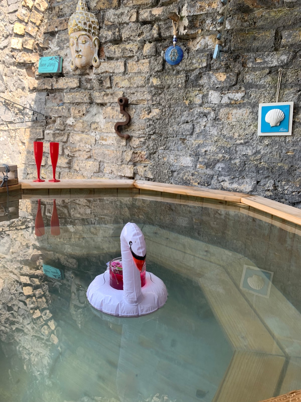 A serene hot tub sits in a private courtyard, surrounded by weathered stone walls. A pink inflatable swan floats atop the clear water, holding drinks. Nearby, two elegant glasses are positioned on the wooden edge, complemented by charming wall decorations, including shells and nautical-themed artwork.