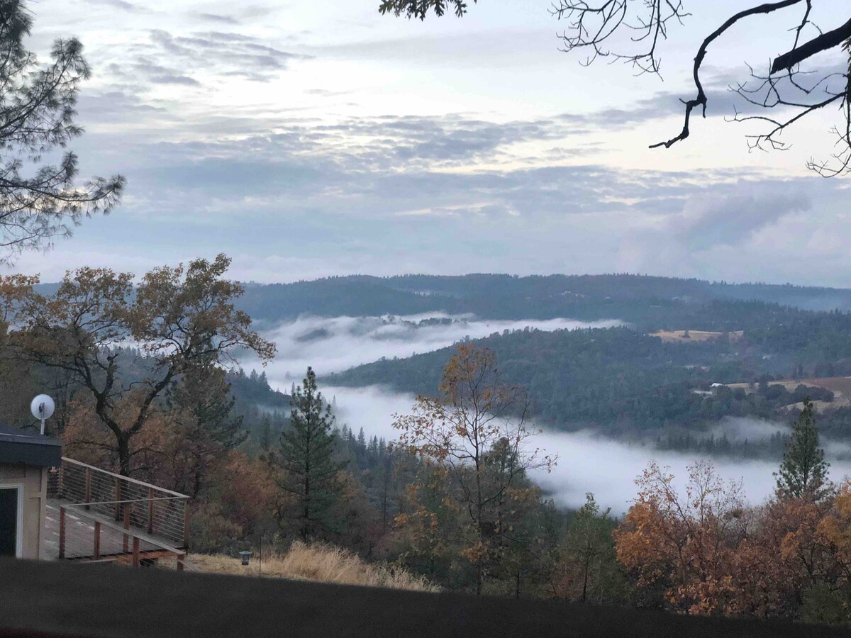 A panoramic view of rolling hills is presented, with mist gently enveloping the valleys below. Trees in various autumn colors dot the landscape, while the sky showcases soft gray tones illuminated by the early morning light.