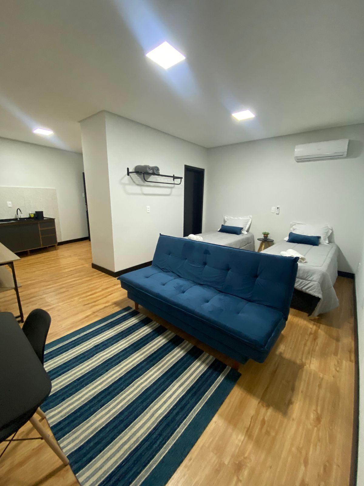 A modern room is equipped with two neatly arranged beds and a blue sofa, providing versatile seating options. Light-colored walls enhance the spacious feel, while warm wood flooring adds a touch of comfort. A small plant decorates the area, contributing to a calming atmosphere.