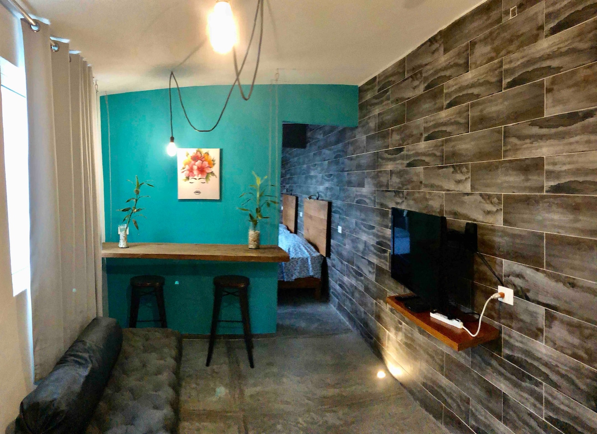 A living area features a dark cushioned sofa and a wooden bar with two stools, complemented by green decorative plants. A wall is adorned with textured gray tiles, while a flat-screen TV is mounted nearby. A painting adds a subtle pop of color to the turquoise backdrop.