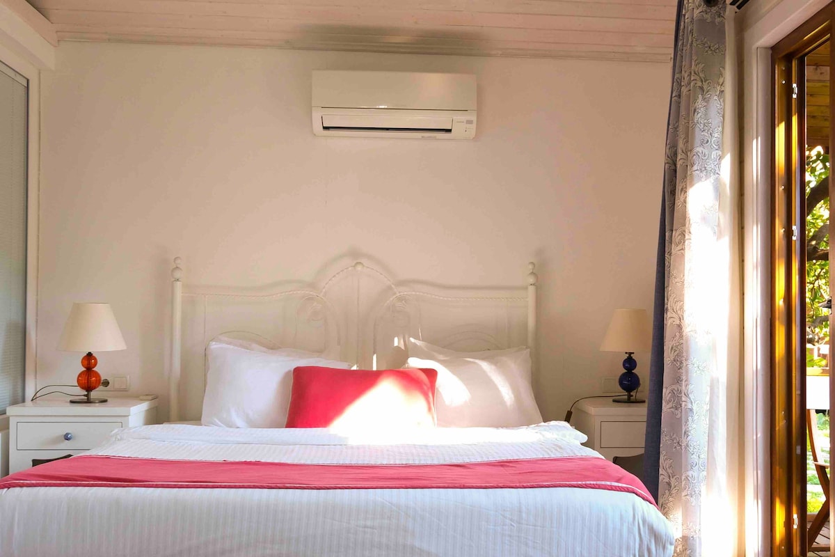 A bright bedroom features a spacious white bed adorned with a colorful throw pillow. Soft lighting is provided by two bedside lamps positioned on matching nightstands. A window allows natural light to enter, and an air conditioning unit is installed above the bed.