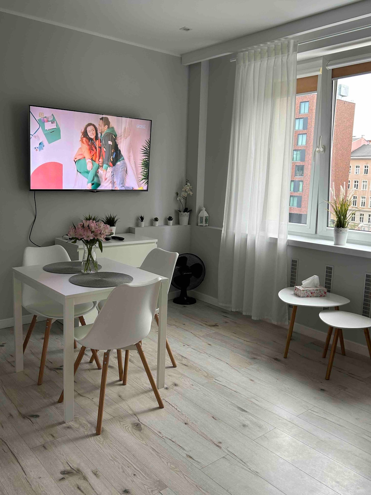 A bright living area is presented with a large flat-screen television mounted on the wall. A white dining table with four chairs is set beside a window draped with sheer curtains, allowing natural light to illuminate the space. Two additional small tables are positioned nearby.
