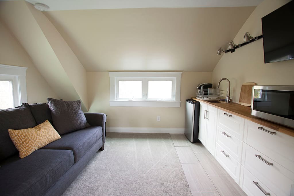 Living room and kitchen. Kitchen is stocked with plates, cups, silverware, coffee pods and tea, cream and sugar, light snacks, and beverages.