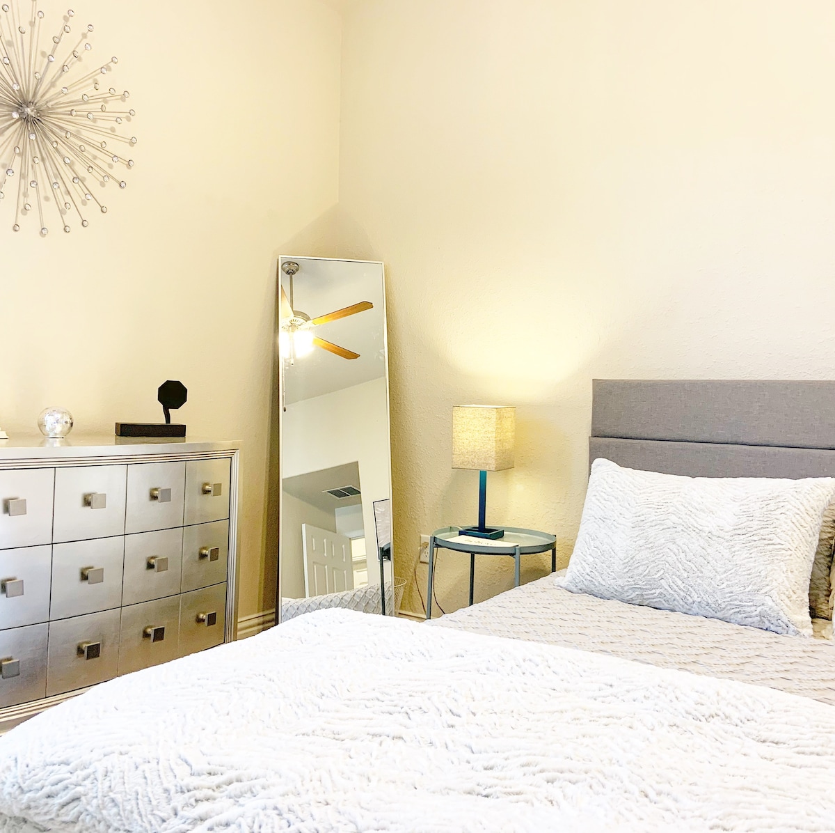 A well-appointed bedroom features a spacious bed with a textured white coverlet. A modern dresser stands against one wall, complemented by a tall mirror and a bedside table with a soft lamp. Light is provided by a ceiling fan, enhancing the room's airy feel.