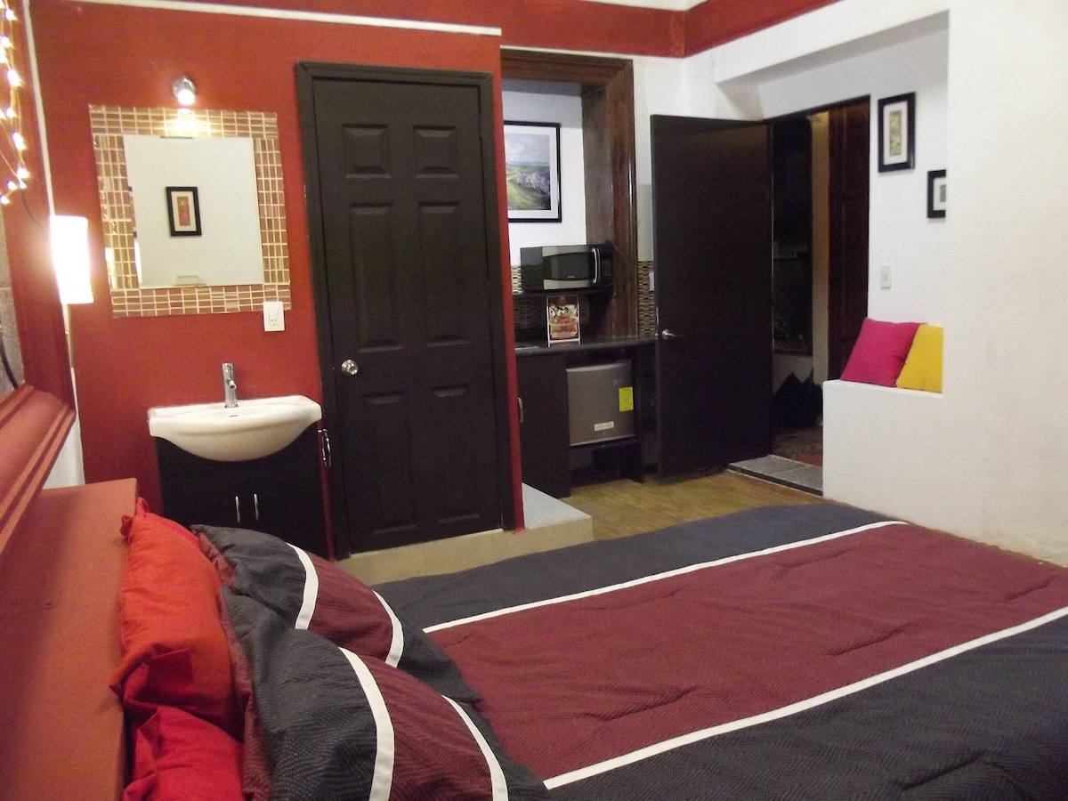 A spacious guest room features a large bed with dark red and navy bedding, complemented by decorative pillows. A small sink is visible near the entrance, alongside a microwave and mini-fridge. The room is framed by deep red walls and soft, warm lighting.