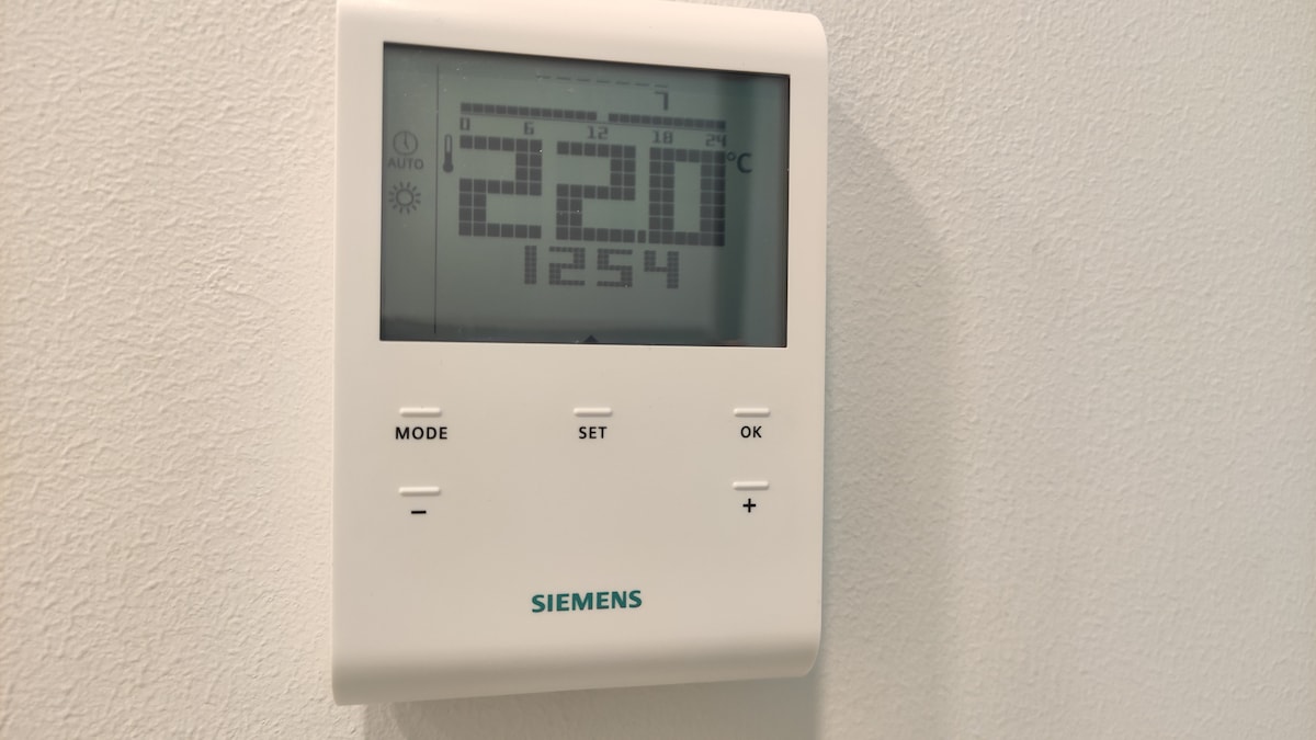 A white digital thermostat is mounted on a textured wall. The display shows the current temperature set to 22 degrees Celsius, with options for adjusting modes and settings clearly visible beneath the screen.