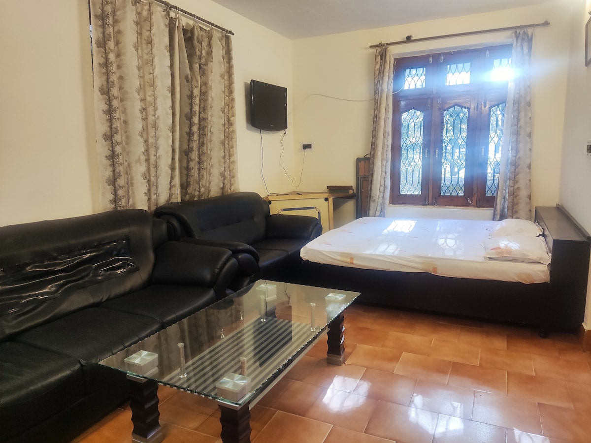 A living area features a dark leather sofa arrangement, complemented by a glass coffee table. A mattress is laid on the floor, adjacent to a window with intricate design allowing natural light. Curtains frame the window, adding texture to the room.
