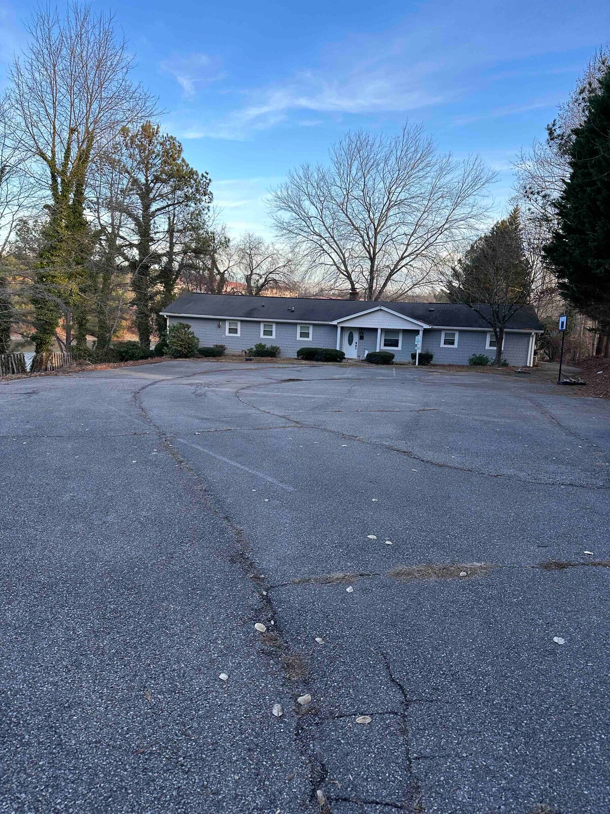 A single-story house is set back from the road, surrounded by trees and a landscaped yard. A spacious gravel parking area is visible in the foreground, with ample room for vehicles. The sky is clear, and the surroundings suggest a peaceful environment.