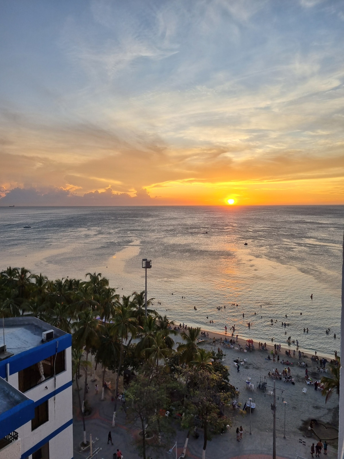 The image captures a vibrant sunset over the sea, casting warm colors across the sky and reflecting on the water. The beach is visible below, with people enjoying the shore and palm trees lining the edge. The scene creates a serene coastal atmosphere.