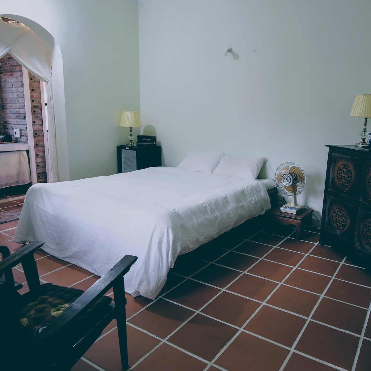 A serene sleeping area features a spacious bed with a white coverlet, set against soft green walls. Two bedside lamps provide gentle illumination, while a ceiling fan adds comfort. A wooden chair and a decorative cabinet enhance the minimalist design of the room.
