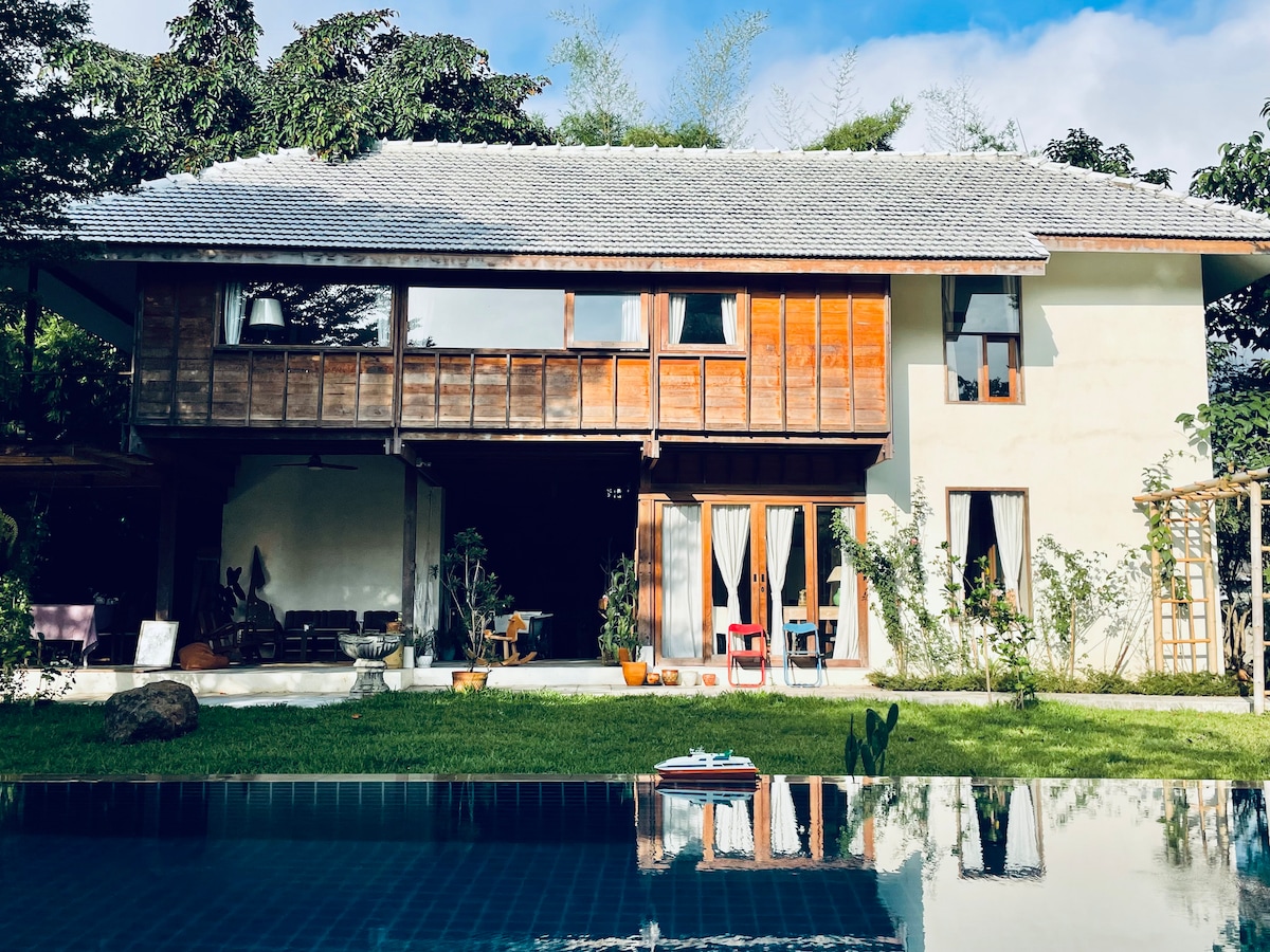 The exterior of a two-story house is depicted, featuring wooden accents and large glass doors that open to the garden. The structure is surrounded by greenery, and a saltwater pool reflects the home and sky. A comfortable seating area is visible on the porch.