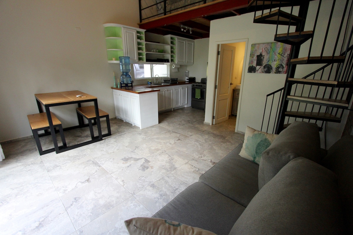 An open living area features a cozy gray couch facing a square dining table with wooden benches. A modern kitchen is visible, displaying white cabinets and green accents. Natural light enters through the large window, illuminating the tiled floor and spiral staircase leading to an upper level.