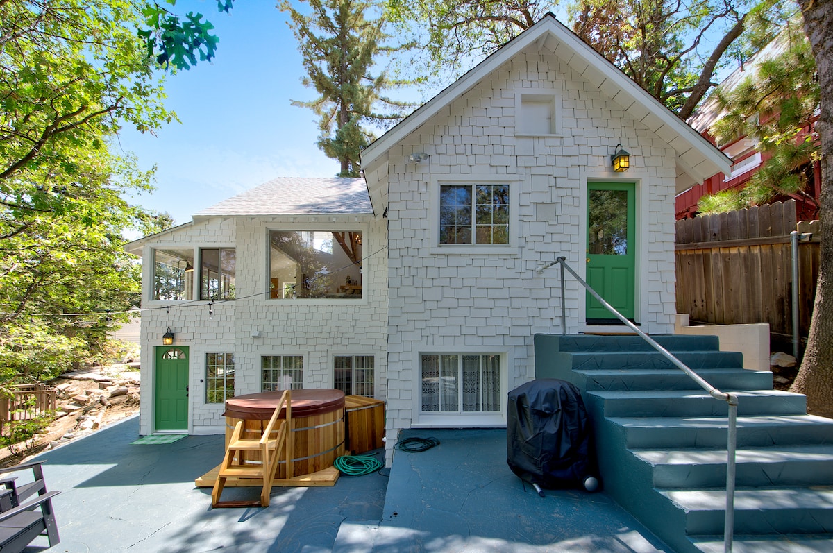 The exterior of the cottage features a unique white shingle design, complemented by green doors and windows. A cedar hot tub and a BBQ grill are situated on a spacious outdoor patio, surrounded by trees and natural scenery. Stairs lead to the main entrance.