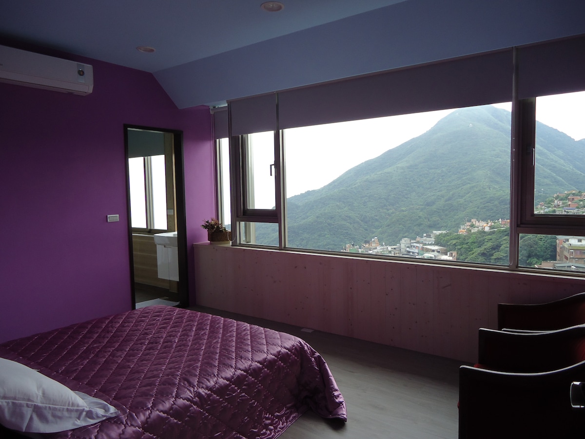 A spacious bedroom features purple walls and a large window showcasing views of the mountains. A quilted blanket in shades of purple covers the bed, complemented by simple wooden chairs nearby. Natural light filters in, creating a serene and bright atmosphere.