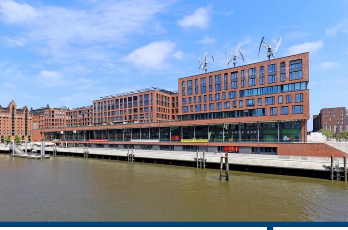 A modern brick building stretches along the waterfront, featuring large windows that frame the view of the water. Wind turbines atop the structure reflect a commitment to sustainability. The gentle ripples of the water enhance the serene setting of the Magdeburger Hafen.