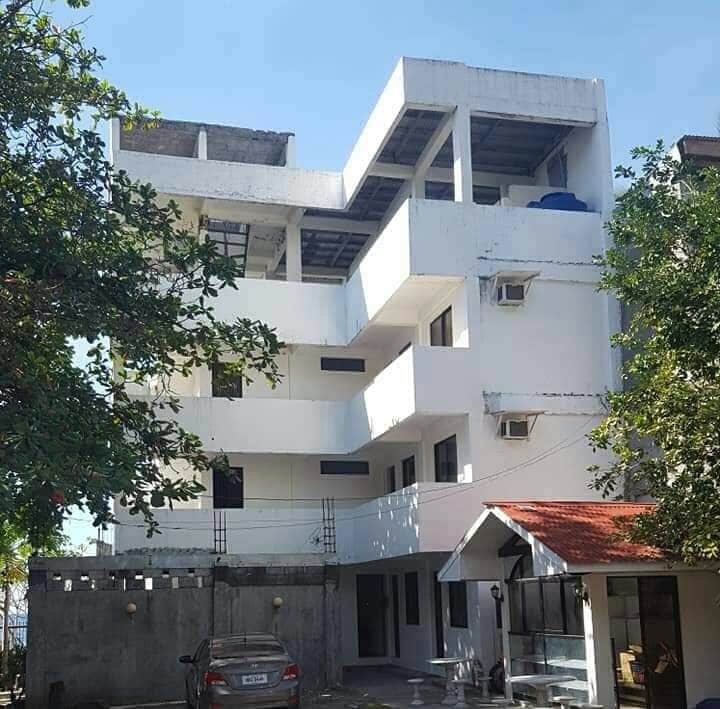 A multi-story building is showcased, featuring a modern architecture design with multiple balconies and large windows. A balcony on the top floor encases an outdoor storage area. A vehicle is parked in the foreground, with green trees providing shade along the pathway.