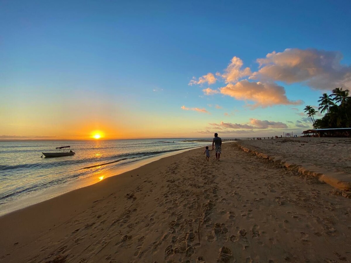 A serene beachscape at sunset features a sandy shoreline, with gentle ocean waves lapping at the shore. A family is seen walking along the water's edge, accompanied by a small boat anchored nearby. The sky is painted with warm hues of orange and soft blue.
