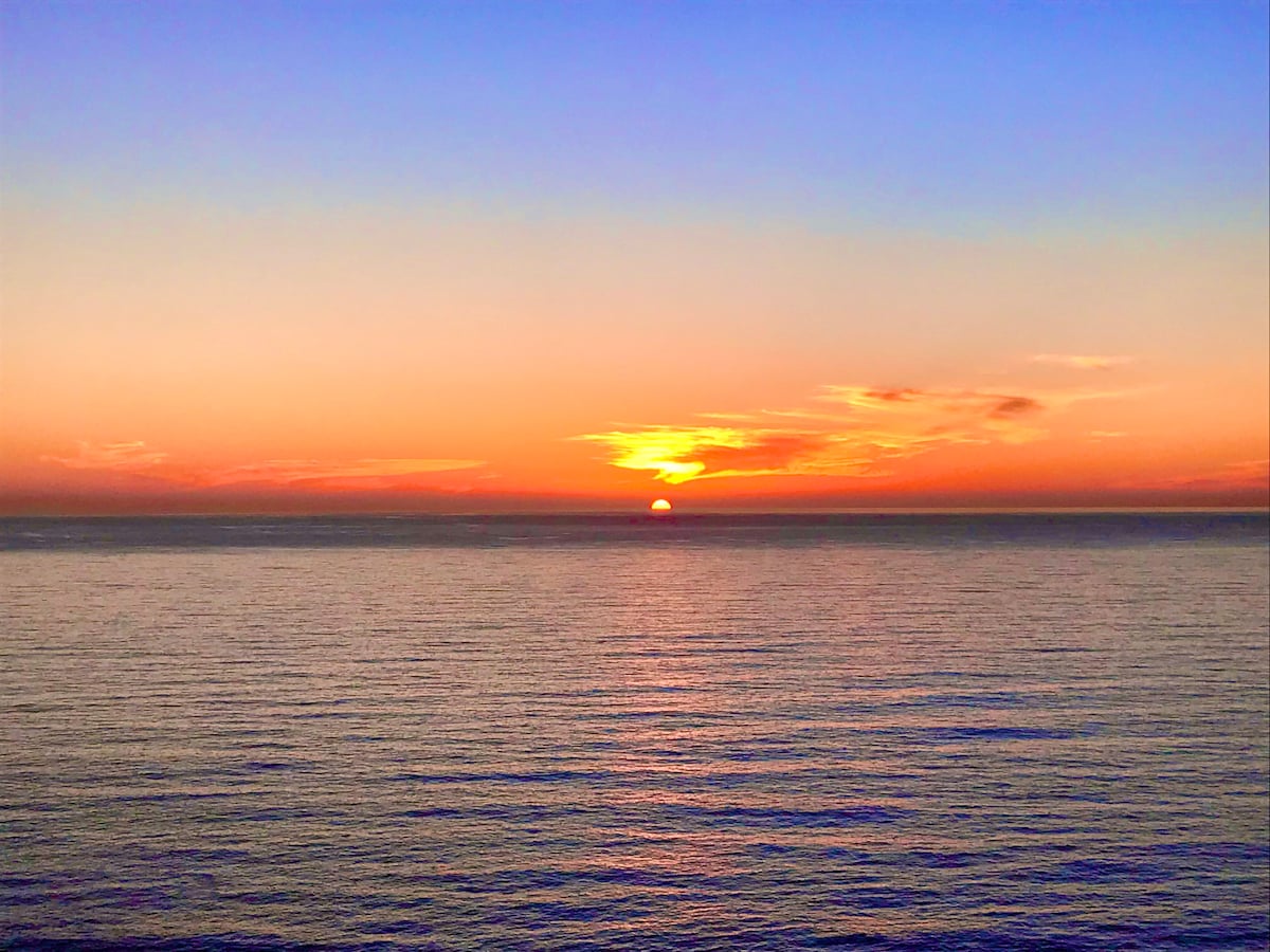 A serene sunset is captured over the calm ocean, displaying a gradient of warm colors from deep oranges to soft purples. The horizon is marked by the sun dipping below, reflecting a gentle shimmer on the water's surface.