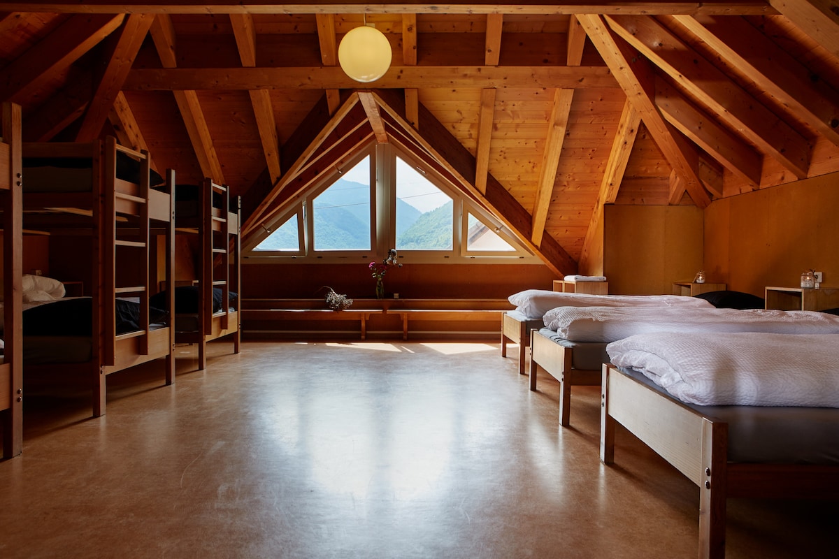 An attic room features wooden beams and large triangular windows offering mountain views. Bunk beds provide multiple sleeping options, while several spacious beds are arranged on the floor. A bench runs along one wall, and light streams in, creating a bright and airy environment.