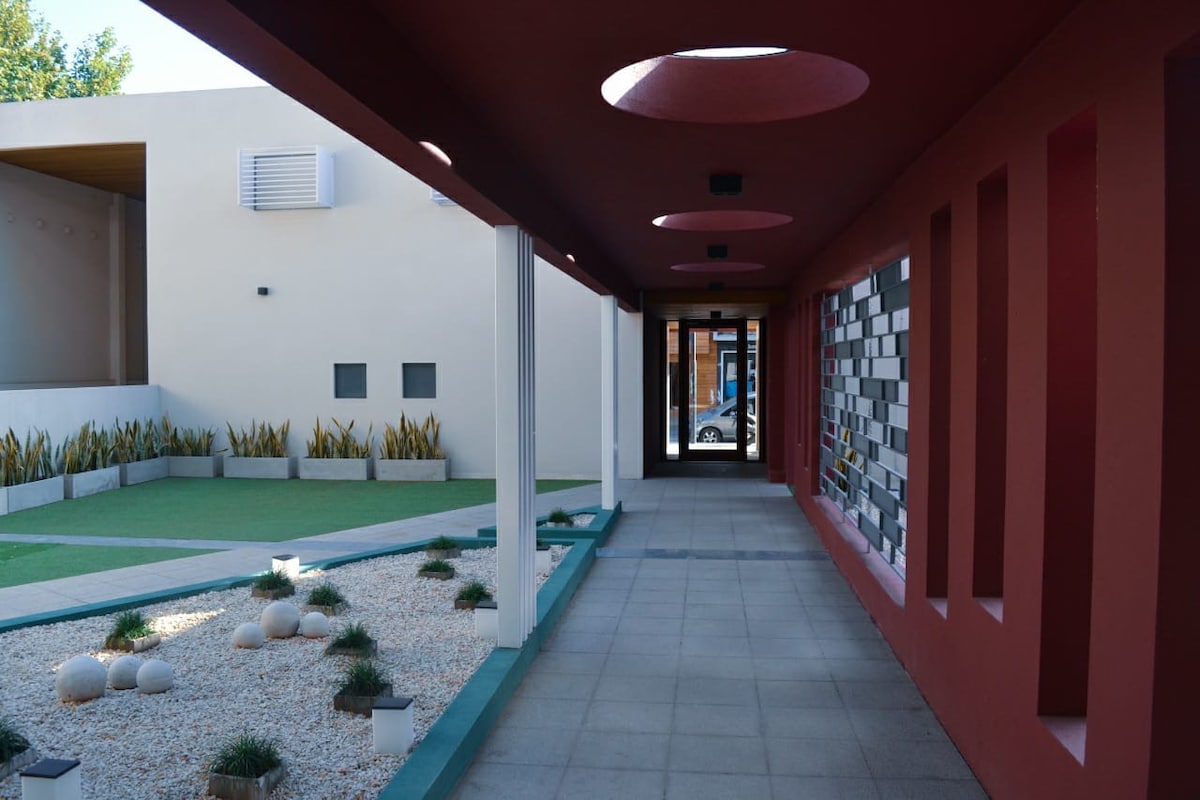 A modern corridor features a minimalist design with a smooth tiled floor. Natural light filters through circular cutouts in the ceiling and the walls showcase alternating patterns of colors. Fresh greenery is visible through expansive windows, enhancing the serene atmosphere within the space.