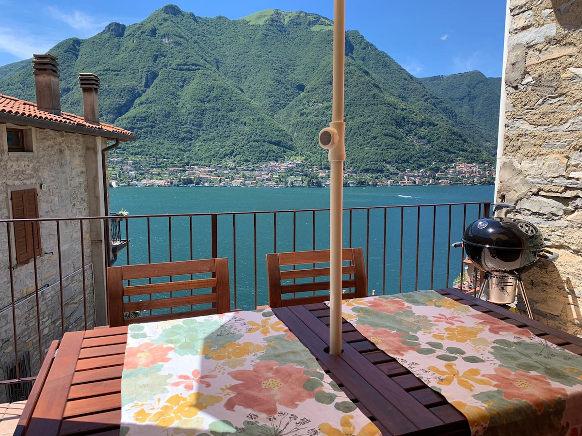 A balcony features a wooden table set with floral-patterned tablecloths, overlooking a serene lake with lush green mountains in the background. A grill is positioned nearby, enhancing the outdoor dining experience.