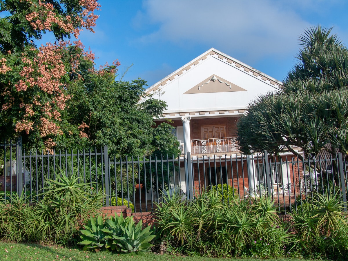 The guest house is set within a lush green landscape featuring various plants and trees. The two-story structure, characterized by a white facade and decorative roofline details, includes wooden front doors and a balcony. A wrought iron fence frames the property, enhancing its charm.