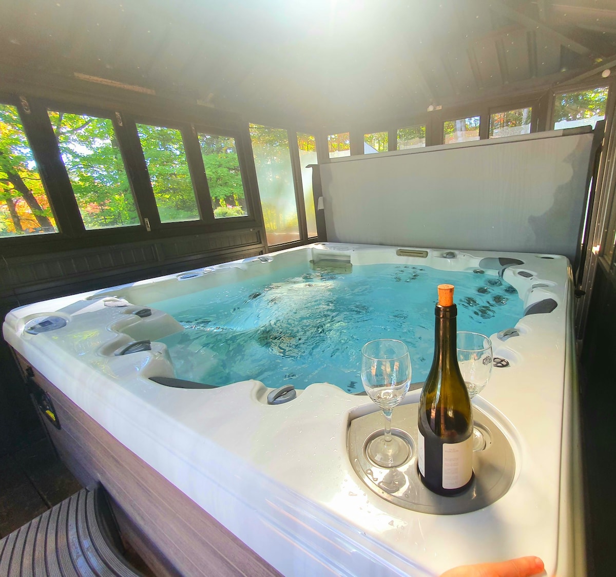 A spacious hot tub is surrounded by large glass windows, allowing natural light to fill the area. A bottle of wine and two glasses are placed on the side, indicating a space ideal for relaxation and unwinding.
