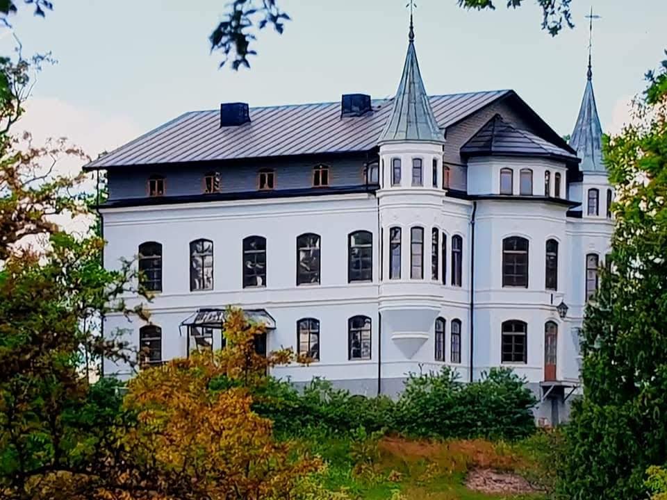 The exterior of Hägerstads Castle is revealed, showcasing its elegant white facade and multiple architectural features. Tall windows and pointed towers are prominent, complemented by greenery at the base. The structure sits on elevated land, framed by trees and natural surroundings.
