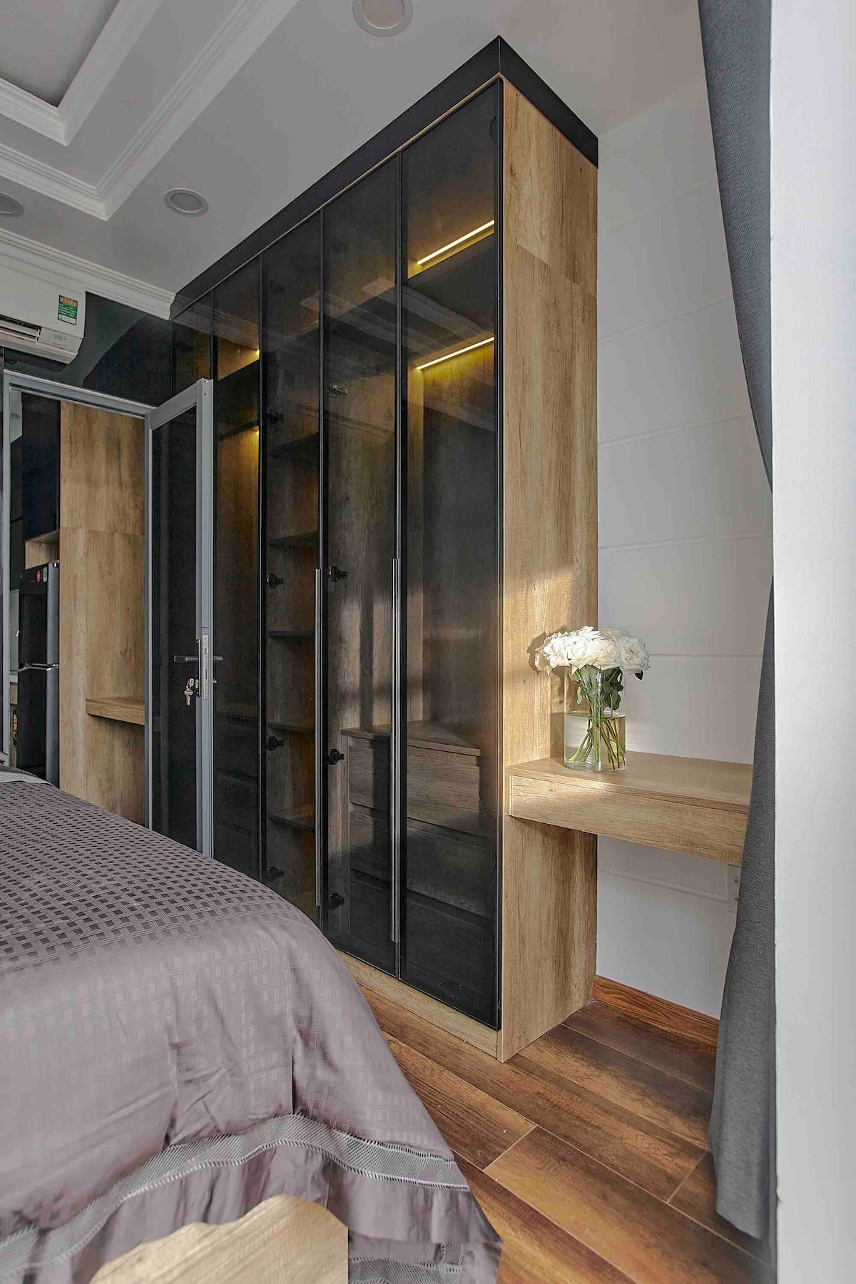 A modern sleeping area features a spacious bed with soft bedding, complemented by a stylish wardrobe with glass doors. Natural light highlights the wooden floors and a small table adorned with fresh flowers, enhancing the clean lines and sleek design of the space.