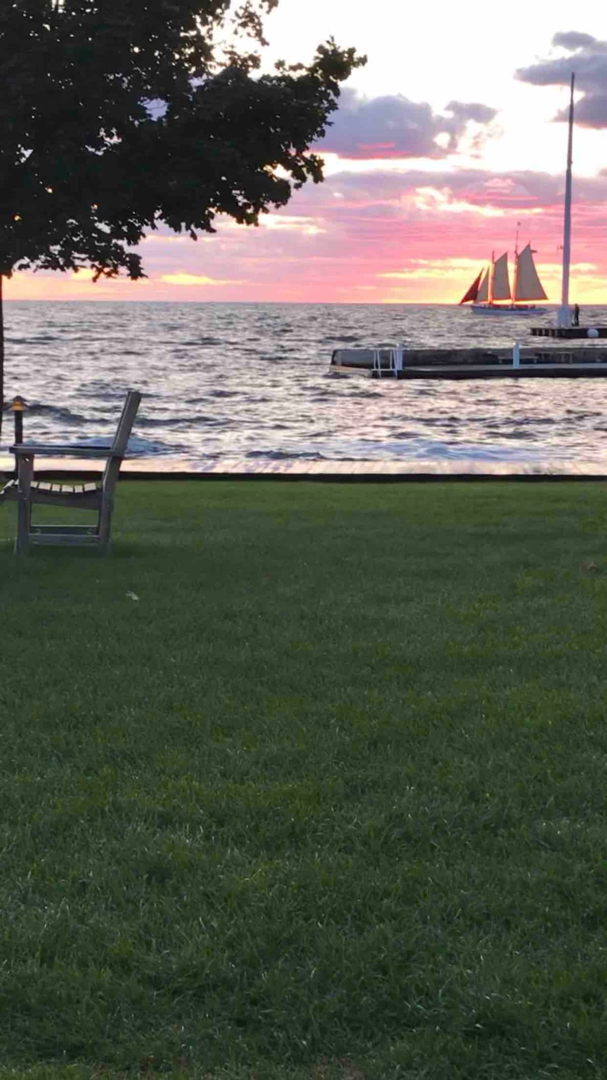 A grassy area is shown with a wooden chair positioned facing the water. The calm bay reflects the soft colors of a sunset, while a sailboat can be seen in the distance near the pier.