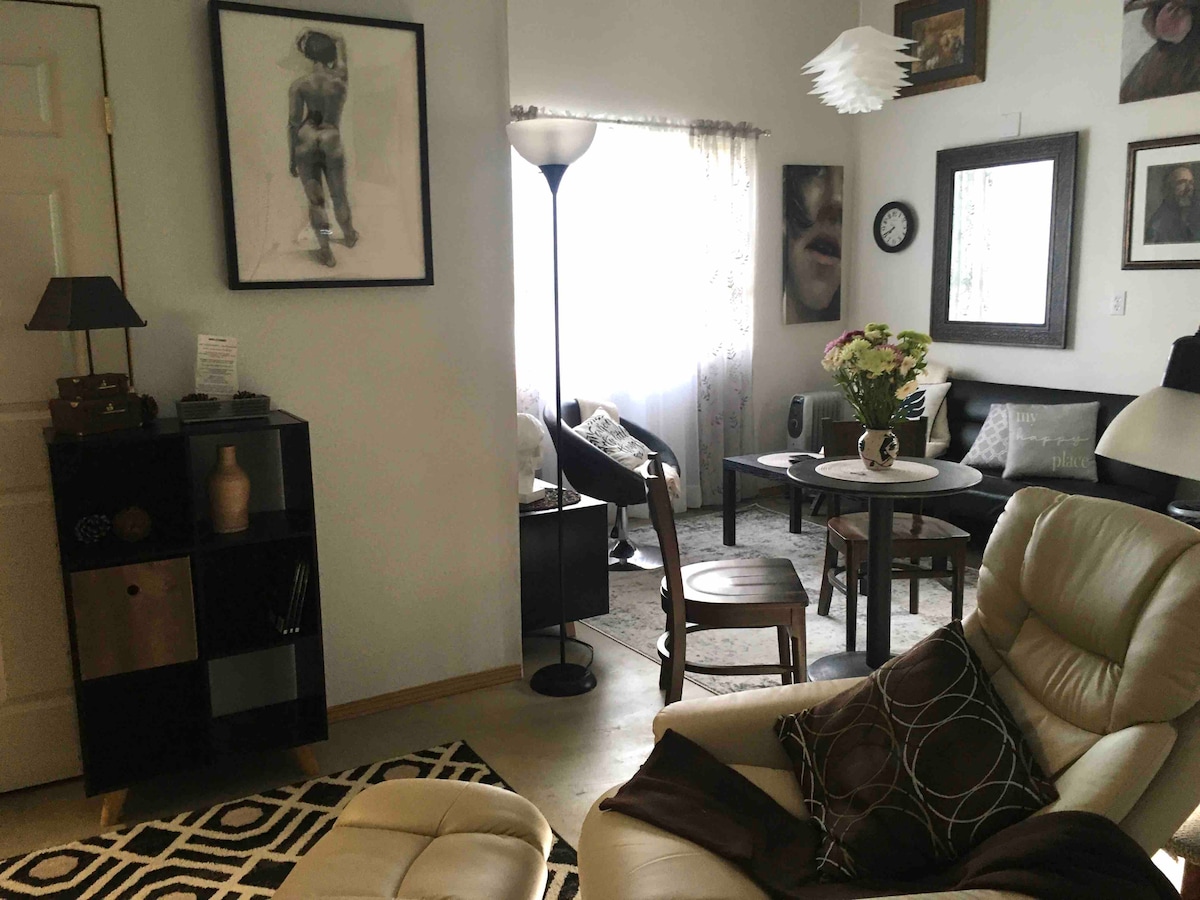 A spacious studio suite is presented with a combination of eclectic art pieces and contemporary furnishings. A comfortable lounge chair and a small dining table are visible, accompanied by a floor lamp providing light. Natural light filters through sheer curtains, enhancing the cozy atmosphere.