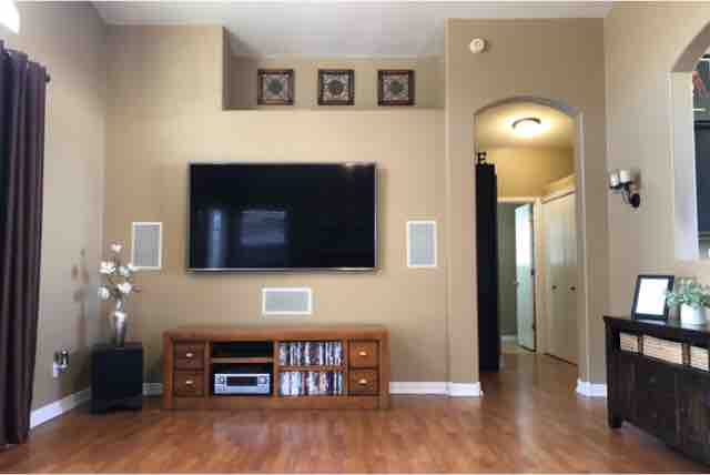 A spacious living area features a large flat-screen television mounted on a light-colored wall. A wooden entertainment center holds media equipment below. Decorative items are arranged on the furniture, while a doorway in the background leads to a hallway. Warm lighting enhances the natural wood flooring.