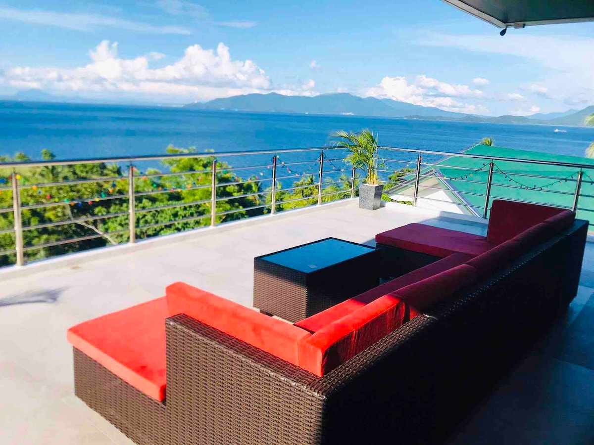An outdoor terrace features a modern seating area with red cushions, surrounded by lush greenery. The space overlooks the expansive ocean, showcasing beautiful views of distant mountains and the clear blue sky. A glass coffee table complements the seating arrangement.
