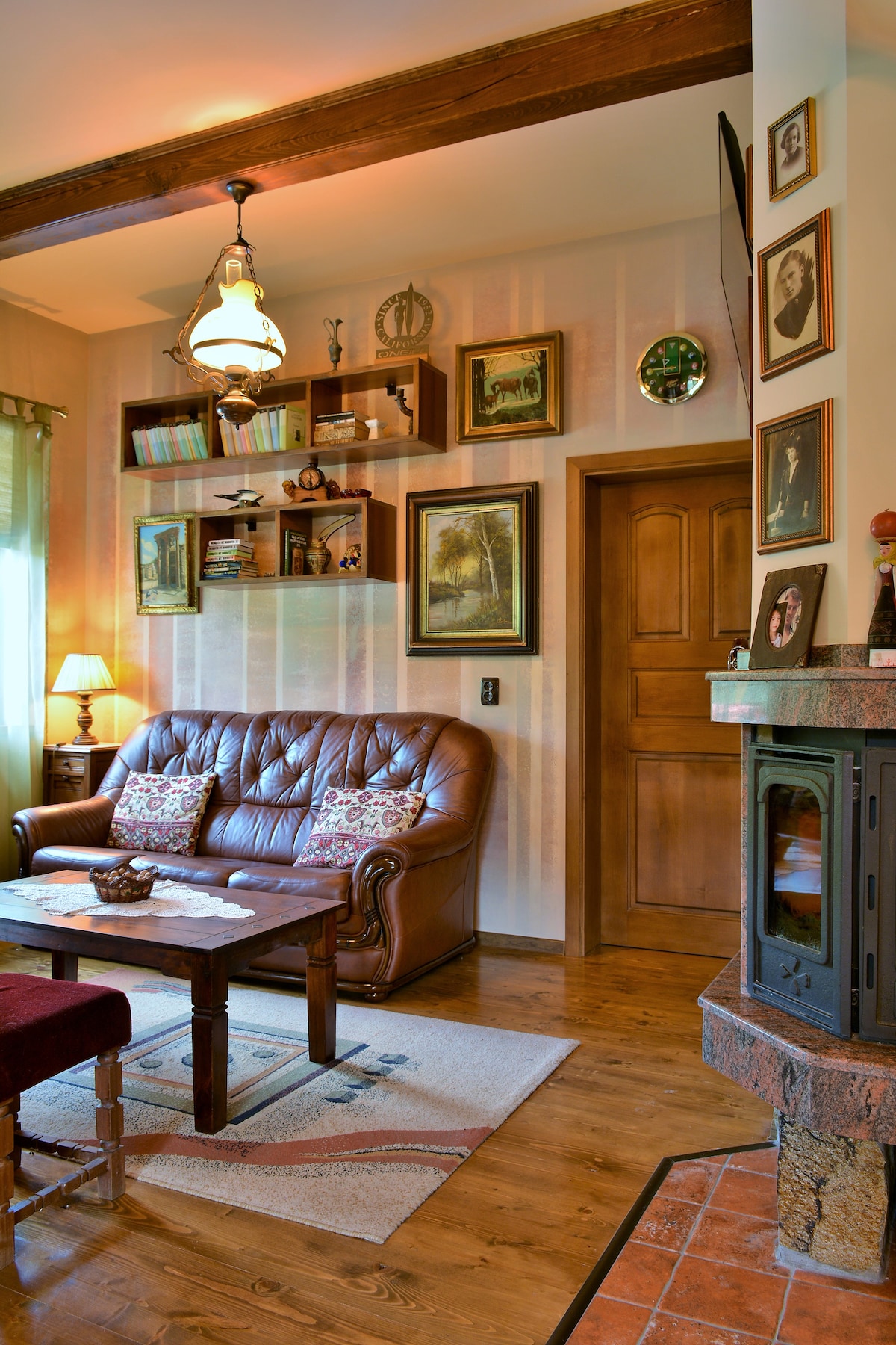 The living room features a leather sofa paired with a wooden coffee table, creating a cozy seating area. Decorative wall shelves hold framed artwork, while a small lamp provides warm lighting. A traditional wood stove adds a rustic touch to the room.