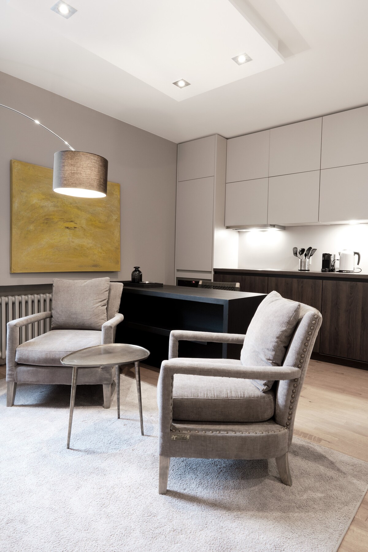 A modern living space is highlighted by two comfortable gray armchairs positioned beside a small round table. Soft lighting from a modern floor lamp illuminates the area, complemented by a large abstract artwork on the wall and sleek cabinetry in the background.