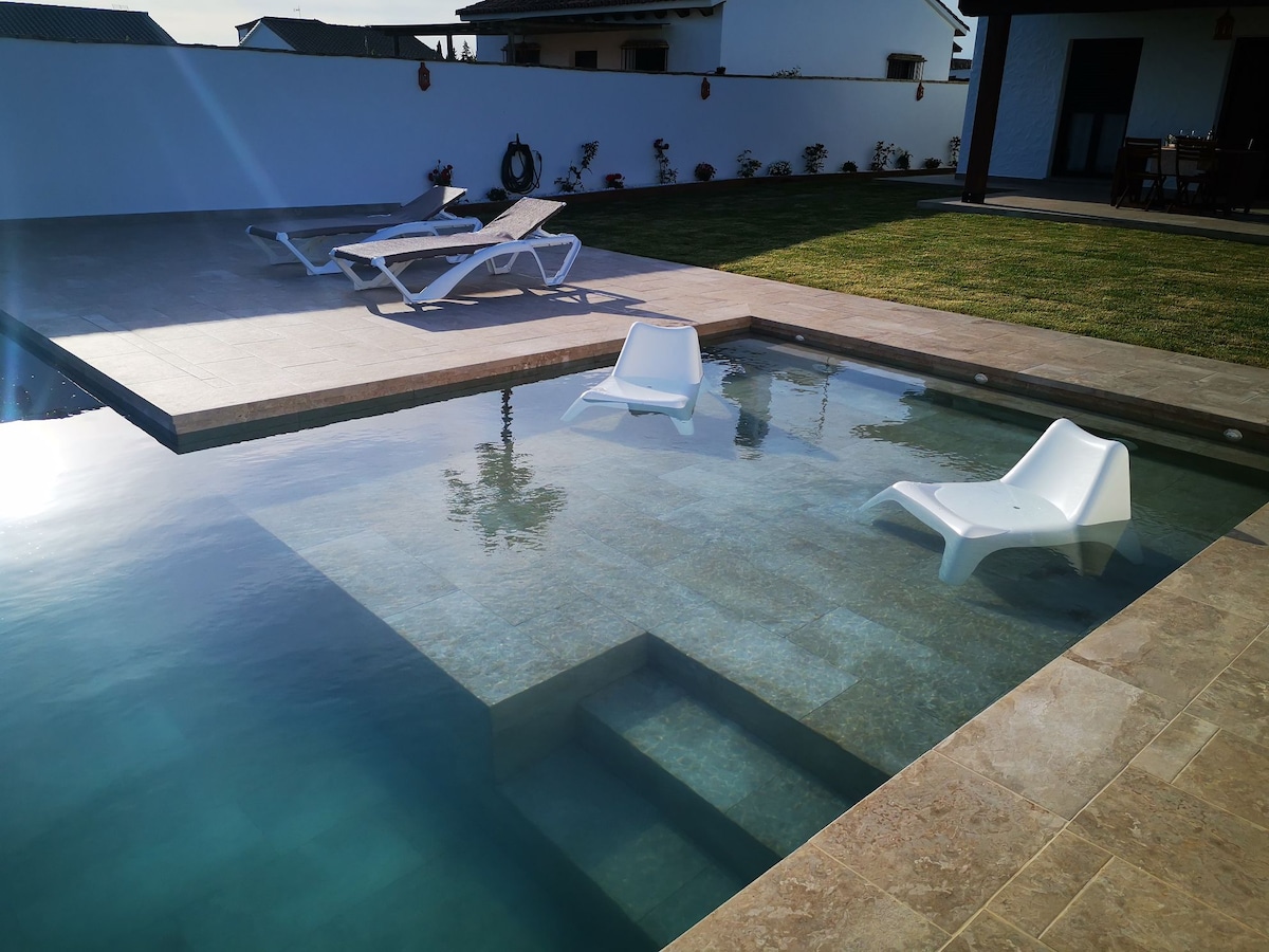 A serene outdoor pool area features two white lounge chairs partially submerged in clear water. Sun loungers are positioned on the surrounding tile deck, providing a relaxing space. A well-maintained lawn frames the pool, contributing to a calm and inviting ambiance.