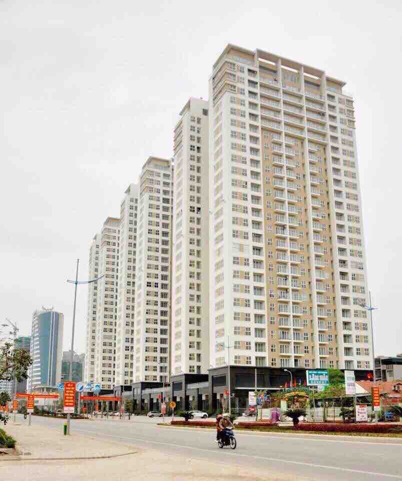 A modern 27-floor building is displayed, featuring a light-colored facade with numerous balconies. The structure is situated on a wide street lined with storefronts and greenery. Multiple vehicles and pedestrians can be seen, illustrating the bustling atmosphere of the area.