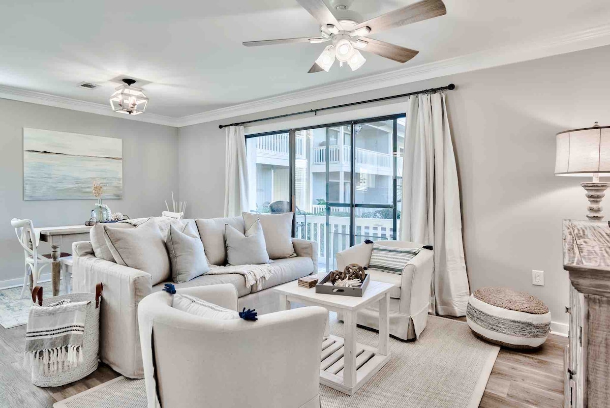 The living area showcases a light-colored sectional sofa, complemented by two armchairs. A coffee table is centrally placed, with decorative items arranged on it. Natural light flows through large sliding glass doors, enhancing a relaxed ambiance. Nearby, a dining table sits with simple styling.