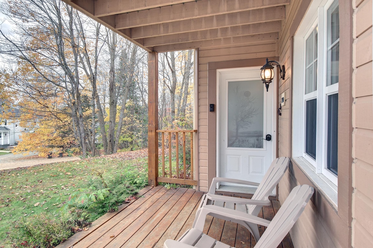 A welcoming porch features two light-colored outdoor chairs positioned on a wooden deck. A door with a frosted glass panel and a lantern light fixture enhance the serene atmosphere. Lush greenery and trees are visible in the foreground, indicating a peaceful natural setting.