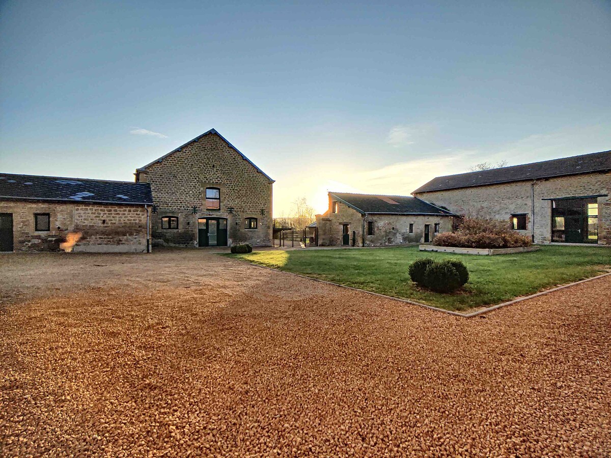 The image captures a spacious courtyard of a renovated farm, surrounded by stone buildings. A gravel pathway leads towards the structures, while an open green area complements the rustic setting. Soft morning light casts a gentle glow, enhancing the serene atmosphere of the surroundings.