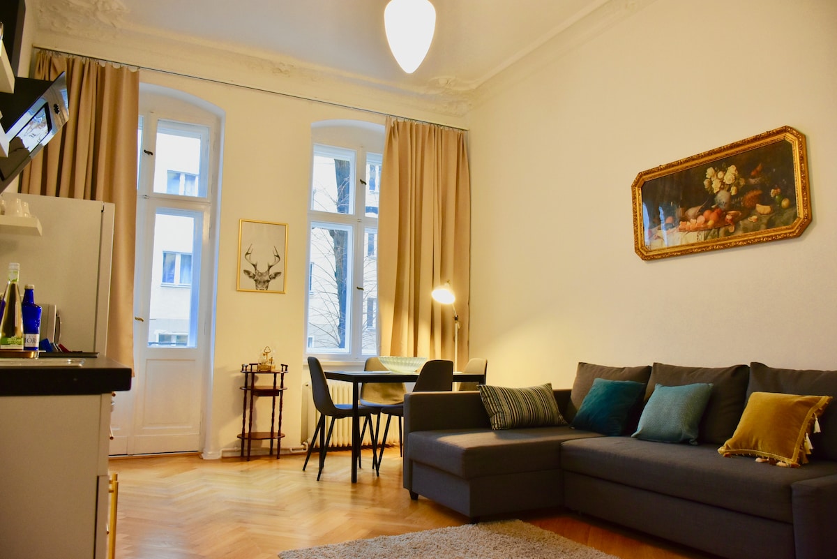 A spacious living area features a comfortable dark gray sofa, accented with decorative pillows. A dining table with chairs is positioned near large windows dressed in beige curtains, allowing natural light to fill the room. Wall art adds a touch of character to the space.